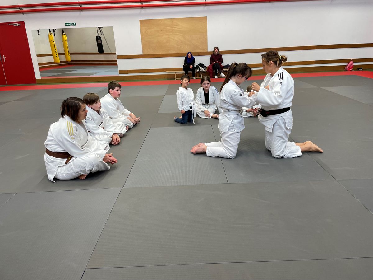 Une professeure noue la ceinture d'une jeune judokate, assises face à face.