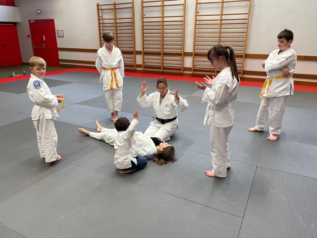 Au dojo, des enfants en judogi lèvent les mains en même temps que leur professeure.