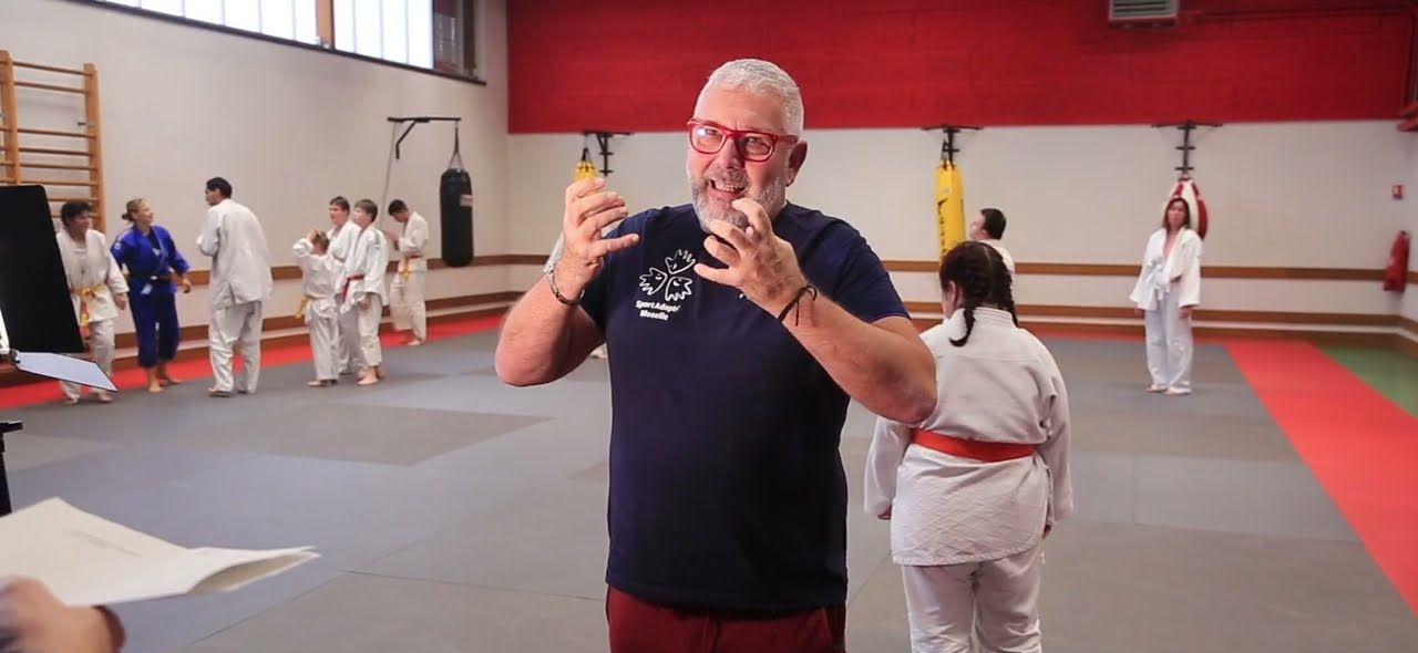 Renato Castellani explique un exercice pendant que des judokas s'entraînent derrière lui.